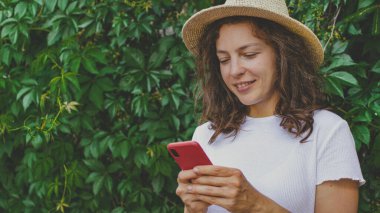 Yazlık hasır şapka ve beyaz tişört giyen akıllı telefon kullanan güzel kıvırcık kız. Sosyal medyayı kontrol etmek, arkadaşlarla sohbet etmek, haberleri okumak, online webinar izlemek, açık havada yeşil bahçede durmak.