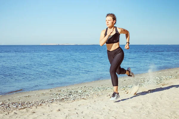 Jogger joven concentrado corriendo en el entrenamiento al aire libre de la playa en la mañana, usando ropa deportiva negra sobre fondo azul del mar y del cielo. Pérdida de peso objetivo cardiovascular desafío logro. Copiar espacio. —  Fotos de Stock