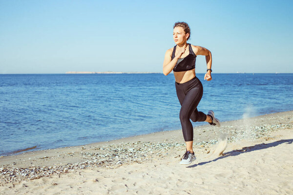 Concentrated young woman jogger running on beach outdoor workout in morning, wearing black sportswear on blue sea and sky background. Weight loss cardio goal achievement challenge. Copy space.