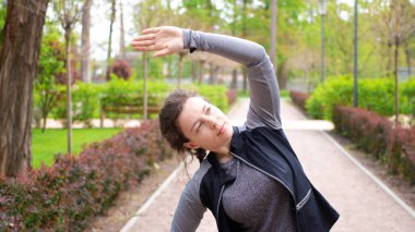 Esmer sporcu kız sabah egzersizi yapıyor koşmadan önce kollarını açıyor ya da yeşil şehir parkında koşuyor. Kolunu yana çeker, dışarıda çalışmadan önce kaslarını ısıtır.