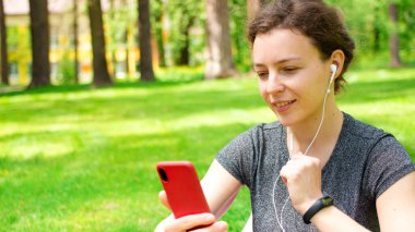Akıllı telefona bakan, şehir parkındaki yeşil çimlerde oturan, kulaklık takan güzel bir genç kadın. İnternette web sitesi, video, dinleme kursu, podcast, sosyal medya izleyen kız öğrenci