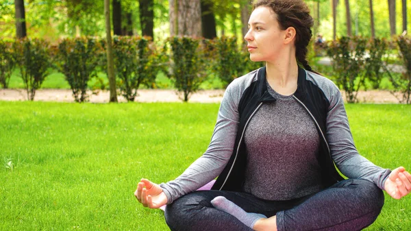 Pretty caucasian girl practicing breathing yoga, sitting in lotus pose on sport mat in park on green grass. Calm female athlete meditating outdoor, looking in distance. Healthy lifestyle. Copy space