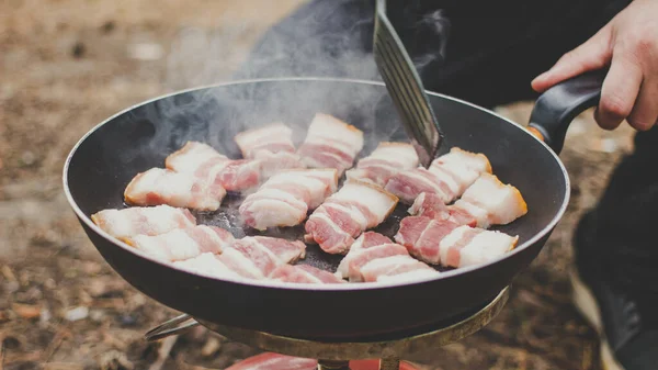 Camping cooking with steam in forest. Man fry bacon in pan on portable gas stove outdoor. Preparing dinner picnic during hike. Touristic campsite lifestyle concept.