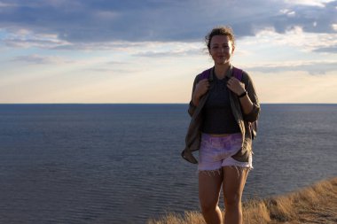 Gün batımında dağın zirvesinde mutlu bir yürüyüşçü kız. Deniz manzarası manzarası. Kameraya bakıp gülümsüyor. Yaz tatilinin tadını çıkarıyorum. Uzayı kopyala Seyahat tutkusu, yeni rota kavramları keşfetmek.