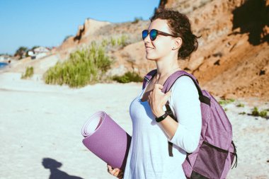 Kumlu sahilde yoga minderi tutan, sırt çantası takan, uzaktan bakan gülümseyen genç bir kadının portresi. Antrenman yapmak için dışarıda beklemek ya da özel antrenör. Beden eğitimi eğitmeni egzersiz geçmişi