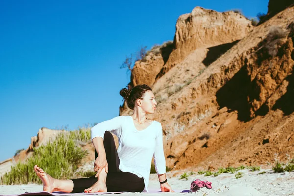 Omurga bükülmesi. Sağlığı için belden aşağısını esneterek yoga yapan sağlıklı bir kadın kumlu sahildeki spor minderinde uçurum manzaralı bir yerde oturuyor. Yaz aylarında temiz hava eğitimi.