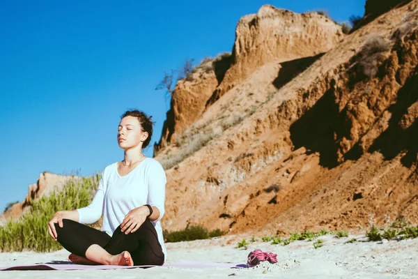 Young calm woman meditating practicing breathing yoga, relaxing sitting in lotus pose with closed eyes on sandy beach with brown mountains landscape view.Training outdoor class. Here and now concept