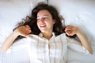 Genç, mutlu esmer kadın, beyaz, rahat bir yatakta uzanmış, gülümsüyor, uzaktan bakıyor, kendini yenilenmiş ve keyifli hissediyor. Yeni bir güne başla, günaydın, uyanma zamanı konsept.