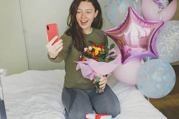 Excited young woman holding flower bouquet using mobile phone for video call sitting in bed at home with colorful balloons. Virtual birthday celebration, surprise delivery. Online congratulations