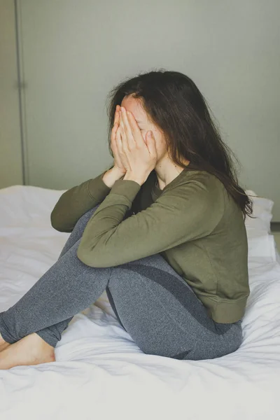 Sad depressed brunette girl covering face with her hands, crying sitting in white bed at home interior. Feeling despair, psychological problem, experiencing grief, relatives death, bad news. Vertical