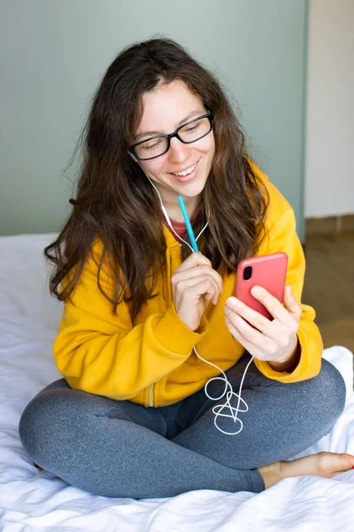 Happy young woman in headphones using smartphone sitting in bed at home. Smiling curly female student watching online webinar,learning language,listening audio course.Online education.Vertical