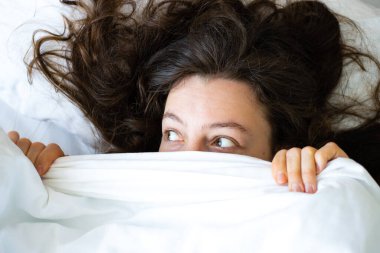 Şaşırmış genç esmer kadın beyaz battaniyenin altında saklanıyor, uzaktan bakıyor, yatakta yatıyor. Yorganı çarşafla örterek dikizlemekten utanıyorum. Üst Manzara. Korkunç rüya, kabus konsepti.