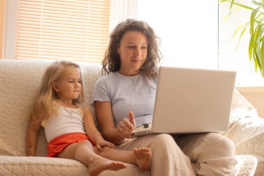 Gülümseyen anne ve evdeki kanepede dizüstü bilgisayar kullanan küçük tatlı kız. Bebek bakıcısı küçük bir kıza bilgisayar kullanmayı öğretiyor. Çizgi film izlemek, video araması yapmak, eğlenmek, web sitesini taramak.