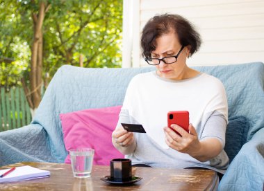 Kredi kartı tutan kafası karışmış yaşlı esmer kadın yanlış anlaşılmaya bakıyor. İnternetten akıllı telefon alıp kanepenin terasında oturmaya çalışıyorum. Borç sorunları, başarısız aktarım, hata 