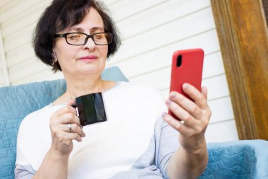 Kıdemli esmer kadın elinde kahve fincanıyla akıllı telefon kullanıyor. Sosyal medyayı kontrol etmek, arkadaşlarla sohbet etmek, haberleri okumak, internetten web sitesi izlemek, evdeki terasta kanepede oturmak. Sabah rutini