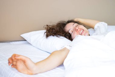 Kalkma vakti. Gözleri kapalı, beyaz örtünün altında uzanmış, sabah uyandıktan sonra kollarını esneten gülümseyen bir kız. İyi uykular. Yatak odasındaki yumuşak yatak ve ortopedik yatağın keyfini çıkarın..
