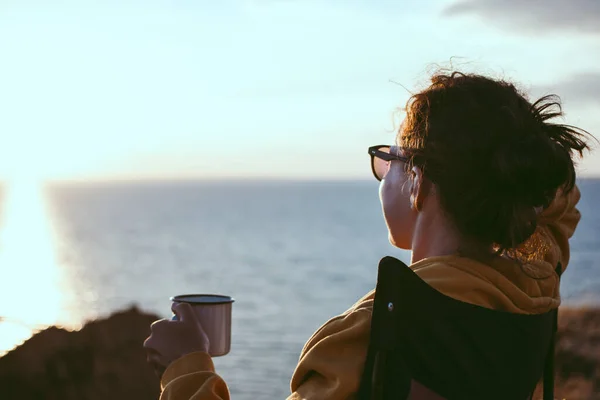 Achteraanzicht van jonge vrouw reiziger met ijzeren beker met thee of koffie, genieten van zonsondergang landschap uitzicht op zee landschap, zitten in toeristische stoel. Reizen camping en avontuur lifestyle concept — Stockfoto