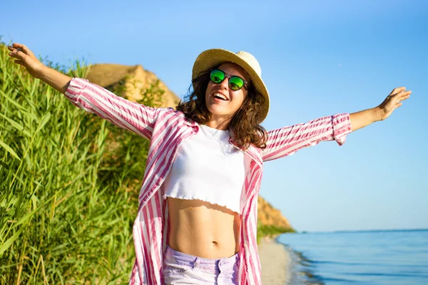 Mutlu, güzel, genç bir kadın yaz şapkası ve güneş gözlükleriyle deniz sahilinde durup gülümsüyor ve uçurum manzarasında cennette hayatın tadını çıkarıyor. Tatil sezonu 2021. Seyahat turizmi