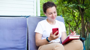 Kulaklıkla gülümseyen, akıllı telefon ekranına bakan, açık terasta oturan genç bir kadın. İnternette web sitesini izleyen, dil öğrenen, ses kurslarını dinleyen, notlar alan bir kız öğrenci.