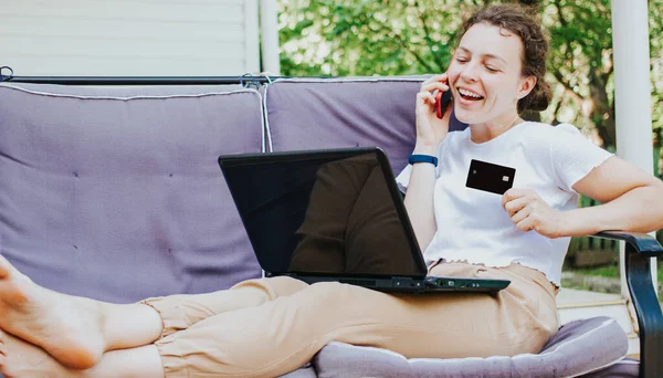 Happy young woman holding bank credit card, looking at laptop, talking on phone with client service, lying on sofa at home terrace outdoors.Making internet payment,online shopping,successful solution — Stock Photo, Image