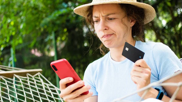 Worried confused young curly woman online buyer holding credit card, looking at mobile phone, sitting on hammock outdoors. Debt problems, insecure online payment, failed transaction, losing money