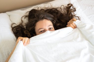 Yatakta uzanmış, yumuşak beyaz yastığı kucaklayan mutlu esmer kadın. Uzaktan gülümsüyor, yatak odasındaki yumuşak yatağın ve ortopedik yatağın tadını çıkarıyor. Rahatlatıcı, dinlendirici, tembel hafta sonu sabahı.