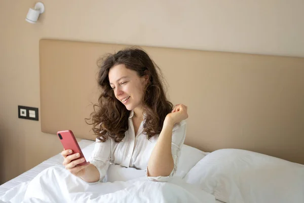 Beautiful brunette young woman looking at smartphone lying in white bed after sleep in morning, using wireless internet connection at home. Chatting with friend, checking social media, messengers.