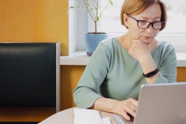 Dizüstü bilgisayarda çalışan son sınıf öğrencisi kadın evin içinde pencerenin yanında oturuyor. Ses kursunu dinlemek, dil öğrenmek, online webinar izlemek, alışveriş, kablosuz internet bağlantısı kullanarak rezervasyon