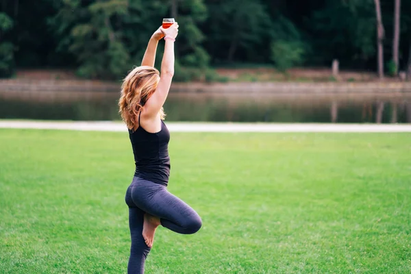 a woman practicing drunk yoga outdoor at the park. Woman holding a glass of alcohol doing pilates. New trending sports classes where people sports with wine or beer or other alcoholic drink to have fun and relax