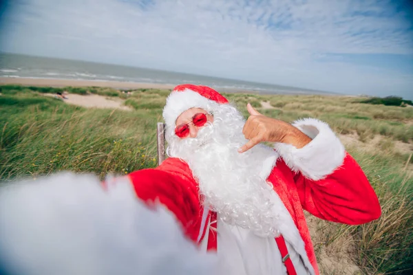 Santa Claus at the sea making a video with his mobile phone while doing the hang loose gesture with his hand
