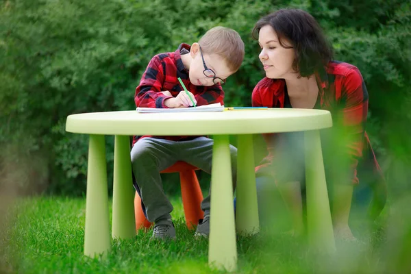 Mom looks at how her little son in glasses draws. Classes with preschoolers in the summer outdoors, in the garden. A boy with a patch over his eye to prevent lazy eye, strabismus, amblyopia.