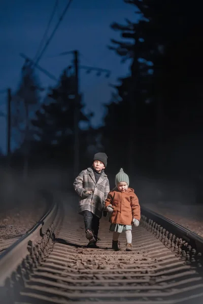 Children walk along the railway tracks at the night. Fog and haze behind. Concept: railroad safety, runaway or lost their parents kids.
