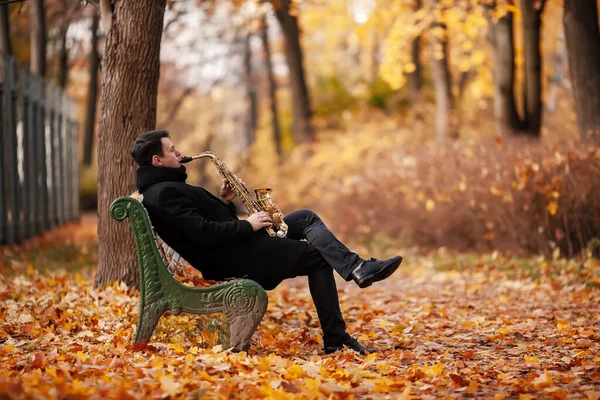 Autumn music: a man saxophonist plays the saxophone sitting on the bench in the autumn park on the background of yellowed foliage.