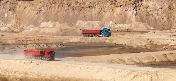 A two dump trucks with semi-trailers ride against the background of a sand pit (quarry, dunes), raising dust behind it. Wide 16:9 image.