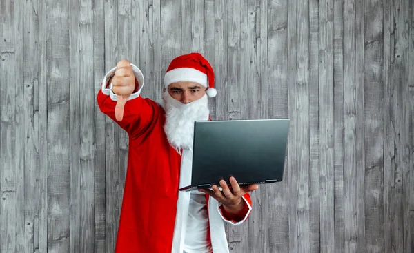 年轻的白人男子穿着圣诞老人的衣服 大姆指朝下 拿着电脑 — 图库照片