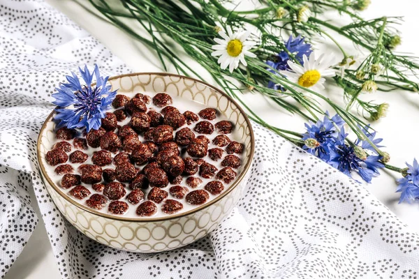 Cereali per la colazione, Cereali al cioccolato nel latte con fiore di fiordaliso su un tovagliolo bianco, concetto di nutrizione sana per i bambini prima di scuola. — Foto Stock