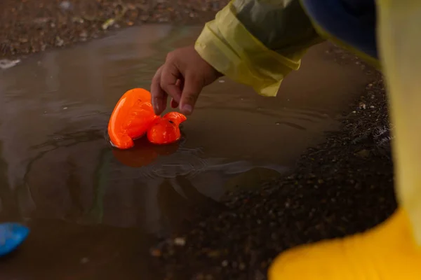 Beautiful girl in denim suit and yellow rubber boots plays with plastic duck and whale in a puddle. High quality photo