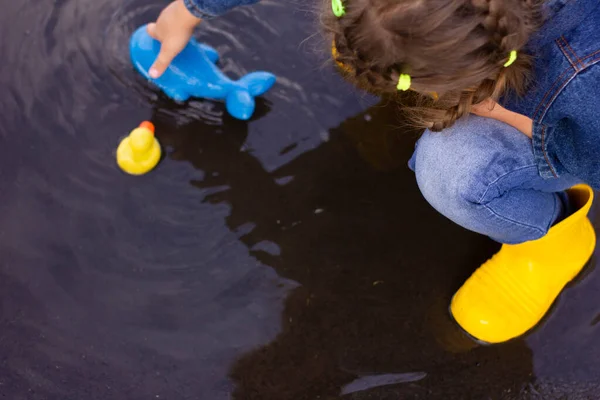 Beautiful girl in denim suit and yellow rubber boots plays with plastic duck and whale in a puddle. High quality photo