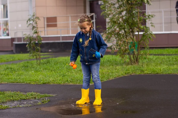 Little beautiful girl in denim suit and rubber boots jumps in a puddle . High quality photo
