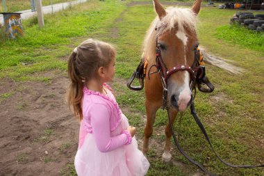 Pembe elbiseli küçük bir kız midillinin yanında duruyor ve onu okşuyor. ipoterapi.