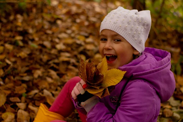 Retrato de uma menina bonita em uma jaqueta roxa e chapéu branco no outono. Ela segura folhas amarelas e senta-se na espuma — Fotografia de Stock