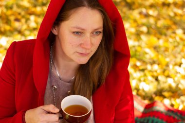 Uzun saçlı, kırmızı paltolu seksi kız sonbahar parkında ekoseye oturmuş ve kahverengi çay içiyor. Yüksek kalite fotoğraf