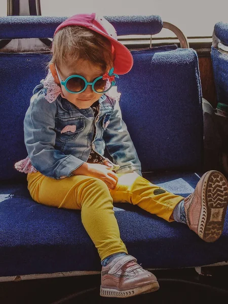 Pequena menina bonita senta-se em uma cadeira no transporte público em calças amarelas e casaco azul e óculos — Fotografia de Stock