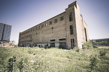 Terk edilmiş, kırmızı tuğla fabrikası. Kırık camlı camlı bir bina. Çimenli bir alanda tek başına duruyor. Renkli bir resim