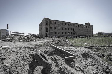 Terk edilmiş, kırmızı tuğla fabrika binası, kırık cam pencereli, enkazla kaplı bir arazide tek başına duruyor. Renkli bir resim
