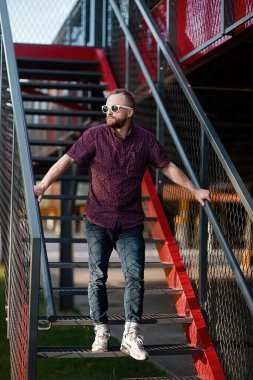 Şık tişörtlü, şık çizgili pantolonlu genç adam şehirde metal bir merdivende oturuyor. Tarz sahibi bir adam yaz günü sokakta rahatlar..