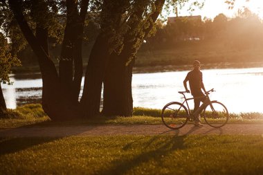 Sağlıklı genç adam bisiklet sürüyor, doğanın ve güneşin tadını çıkarıyor. Özgür bir gezgin gün batımında bir bisikletle seyahat eder. Macera ve seyahat kavramı. Yalnız bisikletçi parkta dinleniyor..