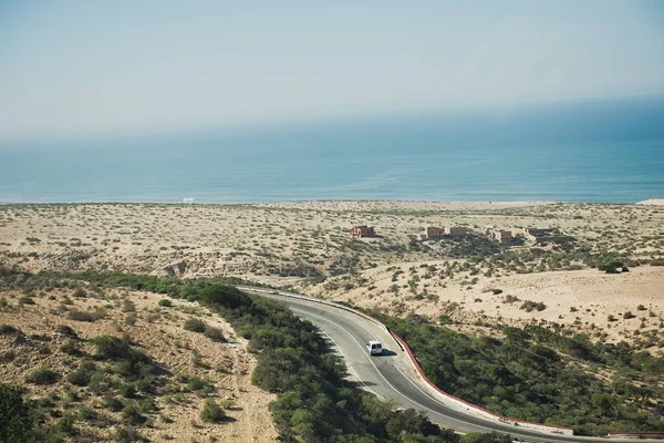 Carro em uma estrada que corre ao longo do oceano a partir de um lado e montanhas no deserto do Saara a partir do outro em Marrocos, — Fotografia de Stock