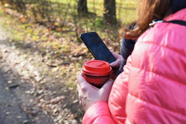 Bir kadın sonbahar parkında yürüyor, akıllı telefon ekranına bakıyor ve kahve içiyor.
