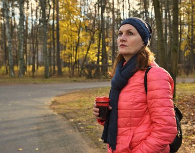 Yetişkin çekici bir kadın sonbahar parkında yürüyor ve kahve içiyor.